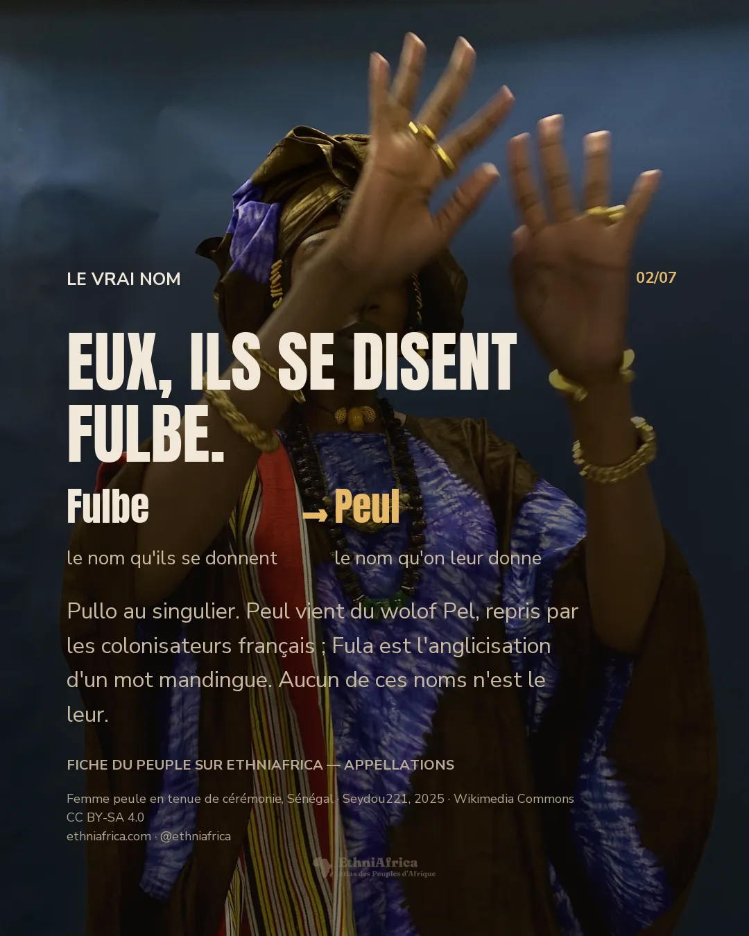 une femme debout devant une toile bleue, les deux mains levées paumes ouvertes devant son visage, en boubou brun sur un tissu teint à l'indigo, bracelets et bagues d'or, colliers de perles et pendentif ciselé