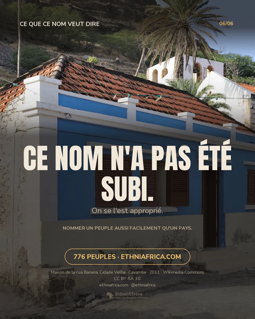 une maison coloniale restaurée, façade bleue à volets bordeaux et toit de tuiles, sur une rue pavée de Cidade Velha, un palmier et une église blanche en arrière-plan, un chien au loin